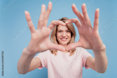 Portrait of cute adorable sweet girl show heart cover face with fingers make frame on blue background