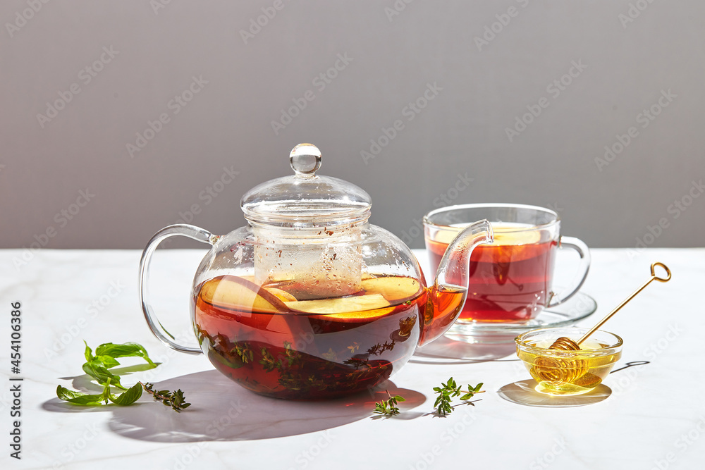 Fruit tea with apples and thyme and honey in glass teapot and cup on white background with hard shadows Fruit tea with apples and thyme and honey in glass teapot and cup on white background with hard shadows