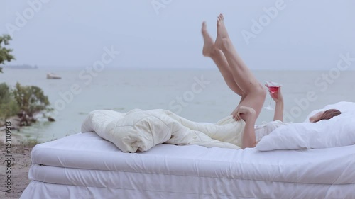 Woman lies on a bed outdoors by the sea. She holds a glass of wine in her hand.