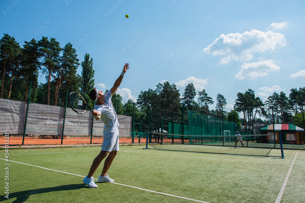 Young sportsman holding racket near ball while playing tennis with african american friend
