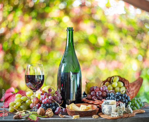 Fototapeta Naklejka Na Ścianę i Meble -  Grapes, bottle of wine and different cheeses on country wooden table and blurred colorful autumn background. Variety of products as the symbol of autumn abundance and prosperity.