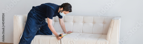 worker in overalls and medical mask measuring white couch in apartment, banner