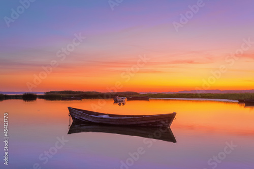 Lonely wooden boat in calm lake. Amazing sunrise in summer morning. The silhouette is reflecting on the water. Orange sky with clouds. Location place Svityaz lake, Ukraine, Europe.