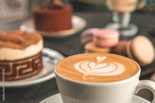 Café con arte latte en un primer plano acompañado de distintos tipos de tortas con foco diferenciado.