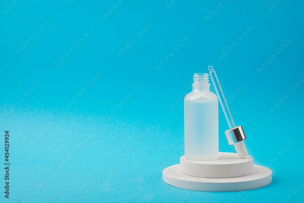 Open frosted transparent glass dropper bottle with serum and a pipette. White podium on blue background. Skincare products , natural cosmetic. Beauty concept for face and body care