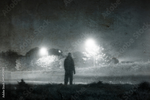 A figure standing by street lights on the edge of town. On a spooky, foggy night. With a grunge retro edit.