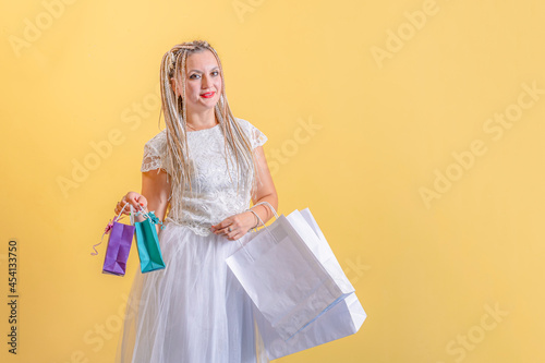 Young woman with African braids on her head, in a white dress, holds bags for purchases and gifts. Stands on a yellow background.