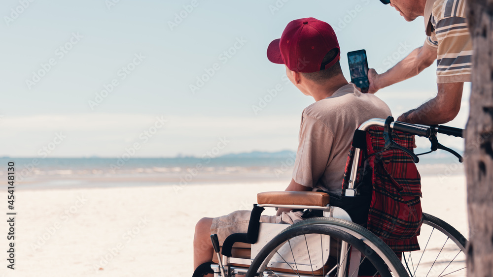 Happy disabled teenage boy on wheelchair and father use social media, Sea beach background, Lifestyle activities in travel of handicapped teen family, Take a selfie and video chat by mobile phone.