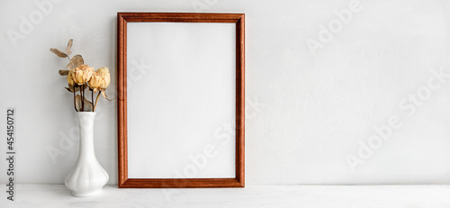 A wooden empty photo frame on a white table and flowers in a vase. A fashionable wedding or holiday banner.