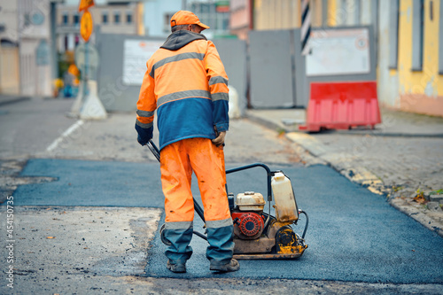 Worker with vibratory plate compactor for road construction. Road repair, worker paving fresh bitumen. Pothole repair, asphalt paving, patching road works with plate compactor.