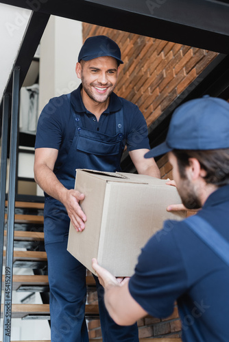 smiling mover carrying carton box with blurred colleague while walking upstairs