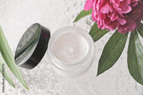 white cosmetic face cream in a glass bottle with black lid. peony flower and shower door. self care and beauty treatment. top view