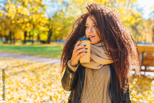 Young afro haird alone woman freezes in autumn park and keeps warm drinking from paper cup