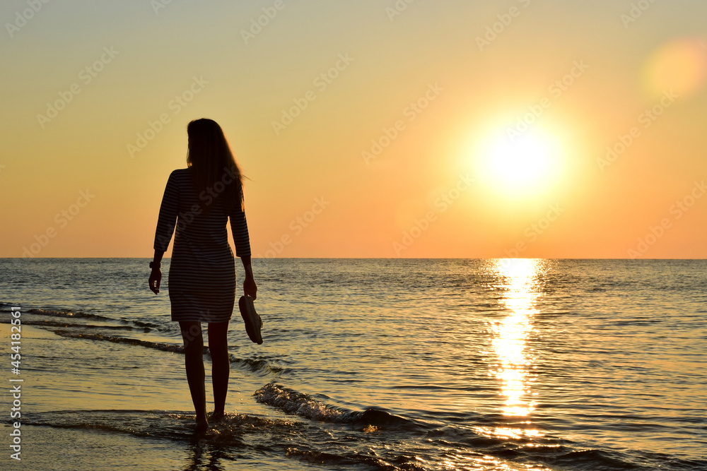 A young blonde girl walks along the beach barefoot and carries shoes in her hand. Back view