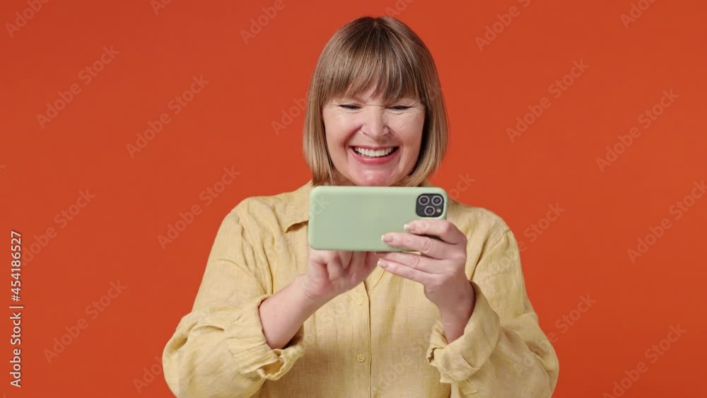 Gambling mature elderly elegant lady woman 55 years old wears yellow shirt using play racing on mobile cell phone hold gadget smartphone for pc video games isolated on plain orange background studio Gambling mature elderly elegant lady woman 55 years old wears yellow shirt using play racing on mobile cell phone hold gadget smartphone for pc video games isolated on plain orange background studio
