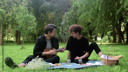 A guy plays the ukulele to another guy in nature in the summer