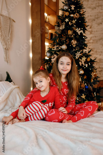 Two sisters in red pajamas are hugging while sitting on the bed against the background of a Christmas tree decorated with toys and garlands. New Year and Christmas holidays with the family
