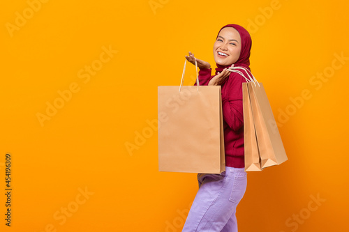 Cheerful beautiful Asian woman holding many shopping bags over yellow background