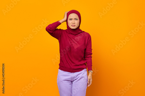 Frustrated confused Asian woman with one hand holding head isolated on yellow background © Bangun Stock Photo