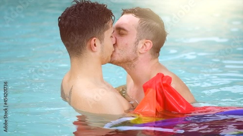 Gay couple relaxing in swimming pool, swimming with LGBT flag. Two young men enjoying nature outdoors, kissing and hugging. Young men romantic family in love. Happiness concept