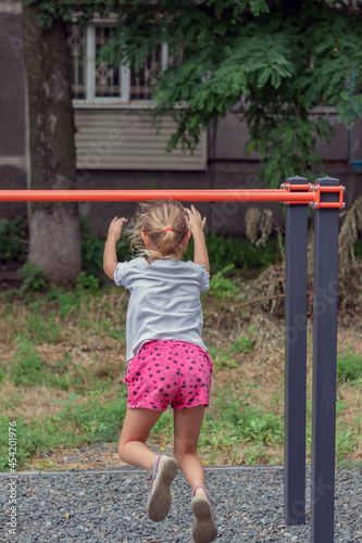 an active child, a girl of 6-7 years old plays on the sports ground, hangs on the uneven bars