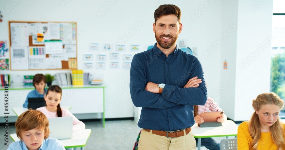 Obraz premium Portrait of handsome young Caucasian male tutor standing in classroom looking at camera and smiling. Children using laptop and tablet studying on background. Happy man teacher at school at work