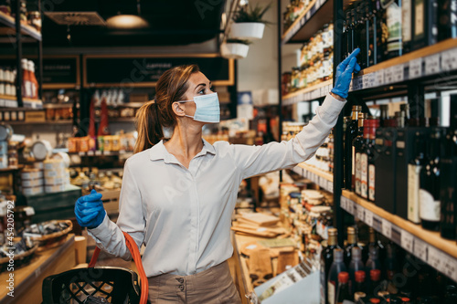 Beautiful young and elegant woman with face protective mask and gloves buying healthy food and drink in a modern supermarket or grocery store. Pandemic or epidemic lifestyle and consumerism concept.