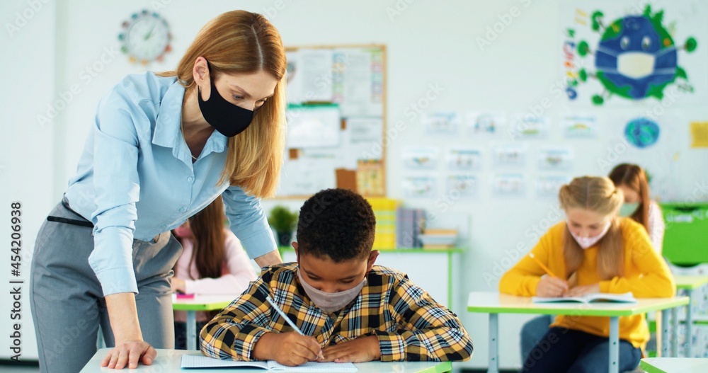 Obraz premium Portrait of young beautiful Caucasian woman teacher helping little African American junior student boy to write test in copybook at class. Female teaching bright schoolboy explaining lesson Post-covid