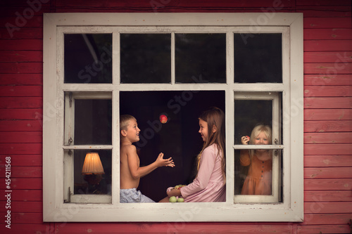 Two cute sisters and funny brother are playing with apples sitting on the old countryside window. Image with selective focus and toning