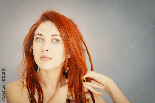 Red-haired girl combs her dreadlocks with fingers. Boho or hippie hairstyle. Woman with pigtails from kanekalon on grey wall background.