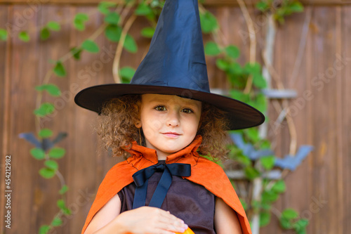 halloween kids. funny cute smiling girl in orange witch costume, black hat with pumpkin Jack, bucket for sweet candy on dark wooden background. scary cheerful autumn holiday for children.