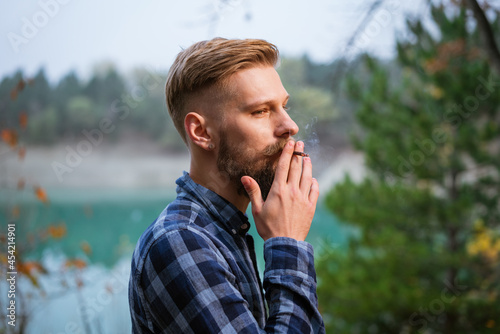 The bearded man smokes. Handsome stylish man in plaid shirt with cigarette. Nicotine addiction and bad habits. Caucasian guy stands in the evening by the lake in the forest © Екатерина Переславце