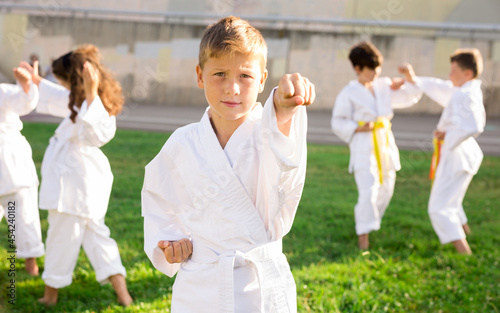 Portrait of positive european schoolboy in kimono practicing karate with his friends in park on sunny day