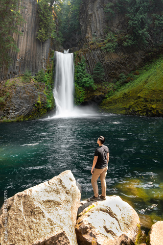 View of a person standing in front of a waterfall in Oregon. Toketee Falls can be seen with cascading water and green moss in a lush forest with a small turquoise pond.