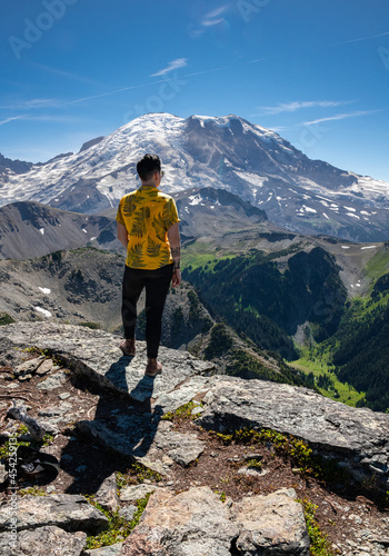 View of a brunette young woman standing in front of Mount Rainier in Mount Rainier National Park. The peak of the mountain is covered in snow and below is fields of green grass.