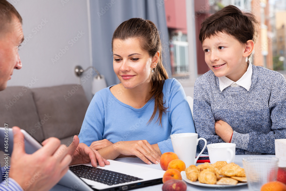 Happy ordinary couple with teenager son using laptop during breakfast at home. High quality photo