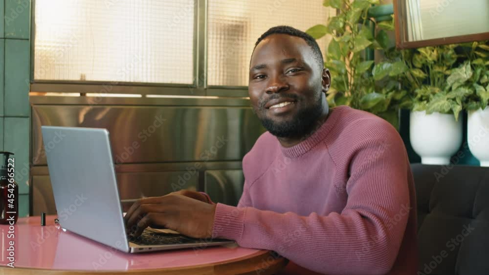 Portrait of young Afro-American man sitting at cafe table with laptop and hand sanitizer on it, looking at camera and smiling Portrait of young Afro-American man sitting at cafe table with laptop and hand sanitizer on it, looking at camera and smiling
