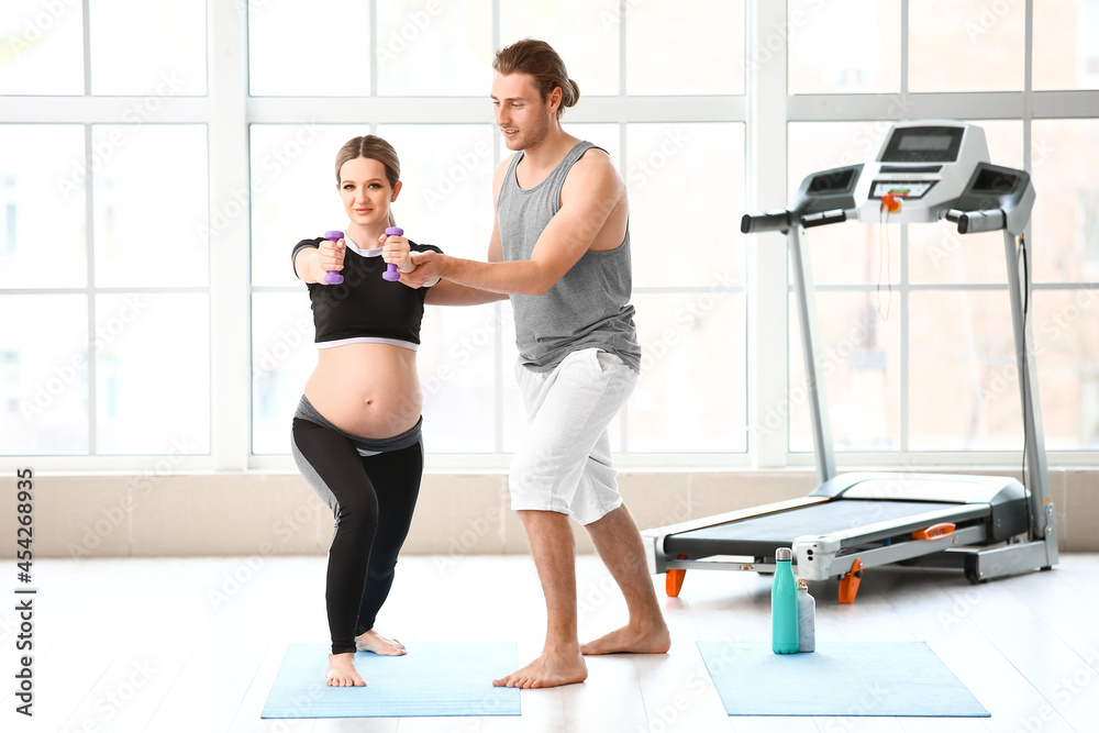Young pregnant woman and her husband with dumbbells in gym