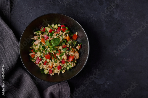 Lebanese traditional salad tabbouleh made of bulgur or couscous, poultry meat, parsley, mint in dark bowl with napkin on black background. Top view, copy space