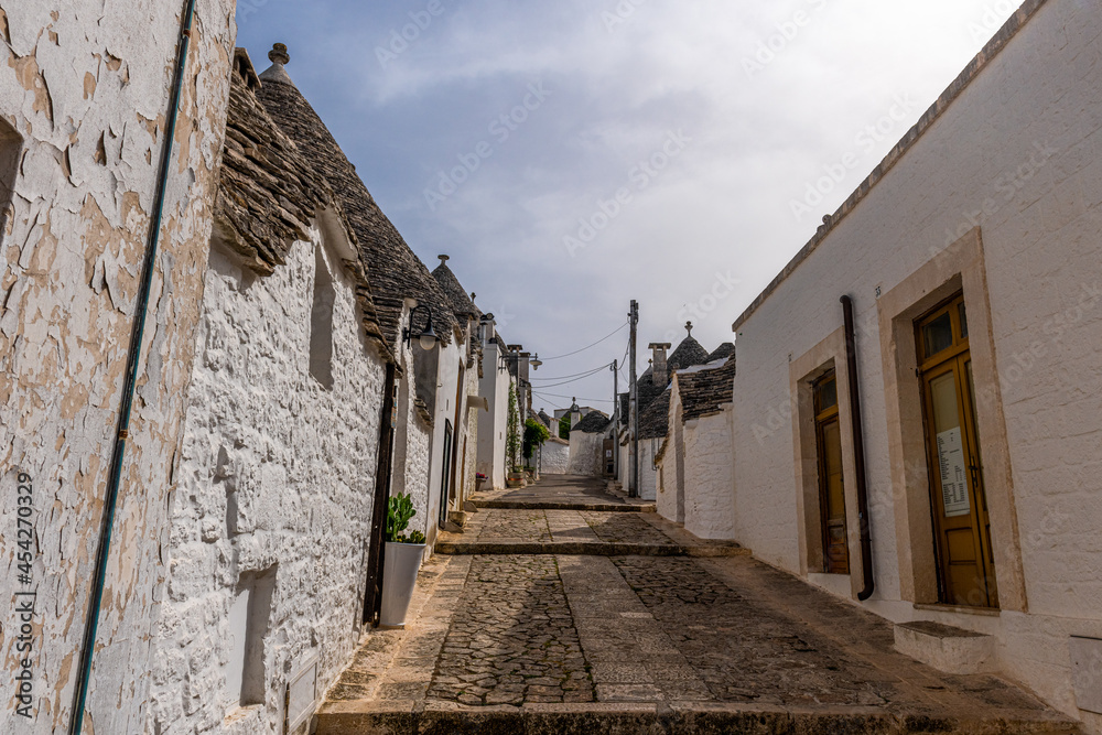 Biegnąca w górę wąska uliczka między Trulli - charakterystycznymi małymi domkami ze stożkowatymi kamiennymi dachami. Alberobello, Puglia, Włochy - obrazy, fototapety, plakaty 