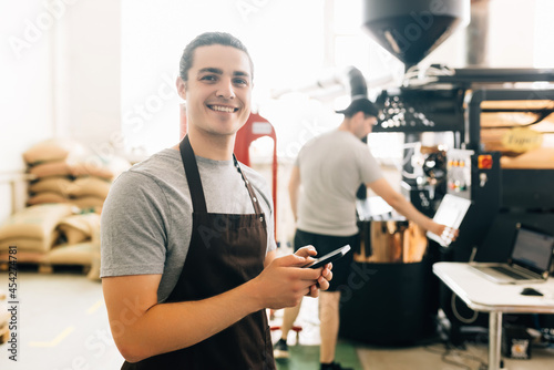 Portrait of handsome modern man wearing apron use phone while standing at coffee roasting machine in local roastery