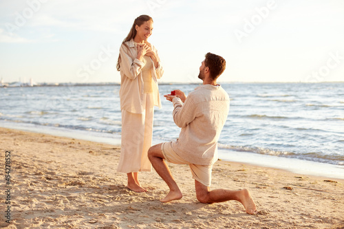 love and people concept - smiling young man with engagement ring making proposal to happy woman on beach