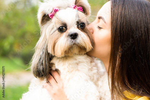 Close up portrait brunette woman holds in her arms hugs and kisses a funny surprised shih tzu dog in autumn park. copy space background. The concept of love and care for animals.