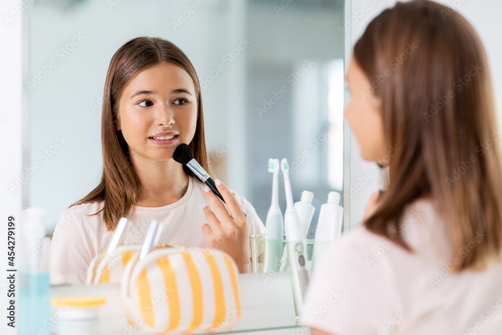 beauty, make up and cosmetics concept - teenage girl with brush applying powder to face and looking to mirror at home bathroom beauty, make up and cosmetics concept - teenage girl with brush applying powder to face and looking to mirror at home bathroom
