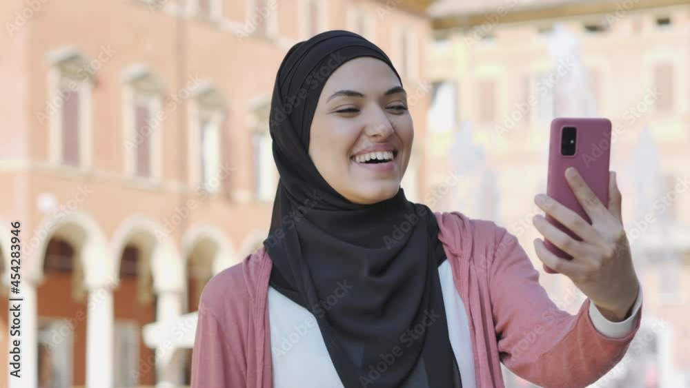 young arab muslim woman in hijab holding mobile phone outdoor having video call chat,smiling middle eastern girl using smartphone waving hand greeting gesture city background young arab muslim woman in hijab holding mobile phone outdoor having video call chat,smiling middle eastern girl using smartphone waving hand greeting gesture city background
