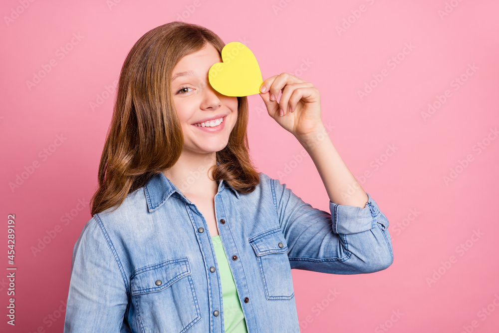 Photo portrait girl smiling hiding eye behind yellow heart postcard isolated pastel pink color background Photo portrait girl smiling hiding eye behind yellow heart postcard isolated pastel pink color background