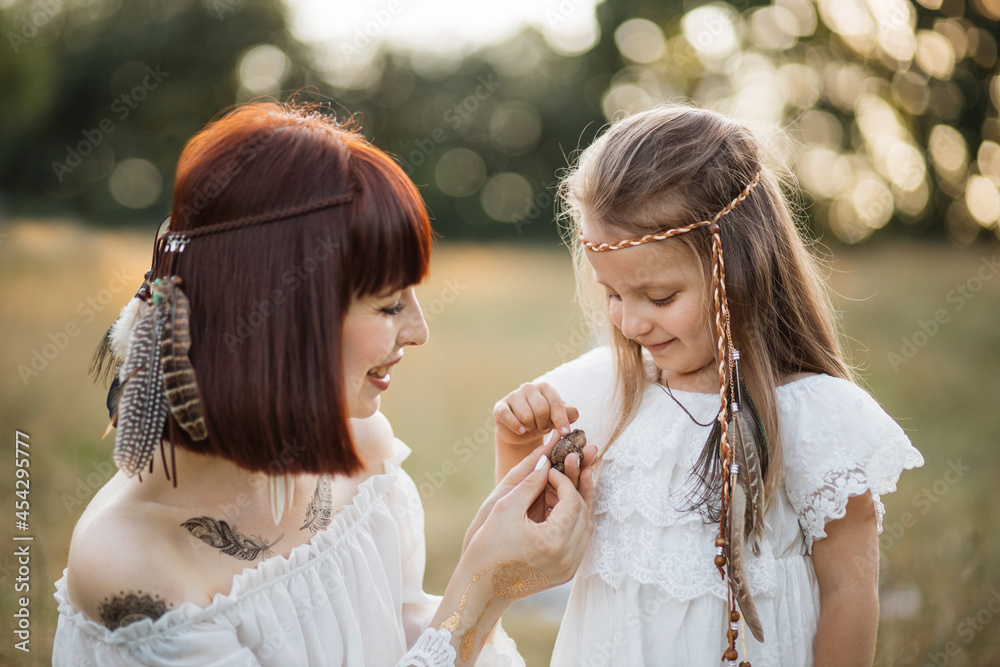 Pretty smiling mother and little adorable toddler daughter in white dress and feathers in hair, playing with little frog in summer field. People and wild animals concept