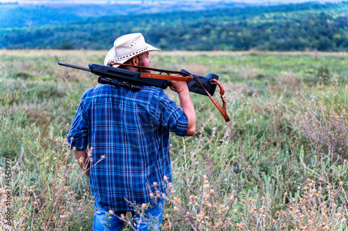 A man with a hat and a rifle on his shoulder stands with his back to the camera and looks to the right, there is little noise, soft focus