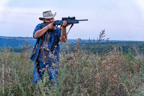 A hunter with a beard and hat shoots a rifle in the field, there is little noise, soft focus