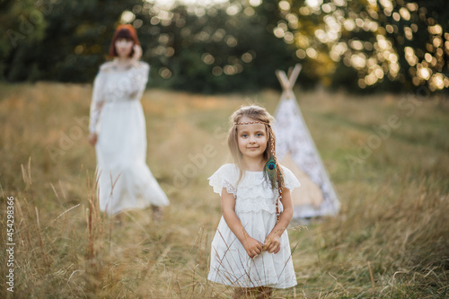 Pretty little blond child girl in white dress and with feather in the hair, posing at camera standing in the summer field at sunset. Woman mother stands on the background near teepee tent